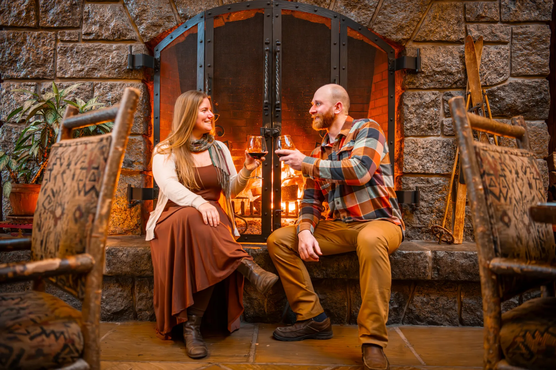 Couple sits in front of stone fireplace with raised glasses of red wine