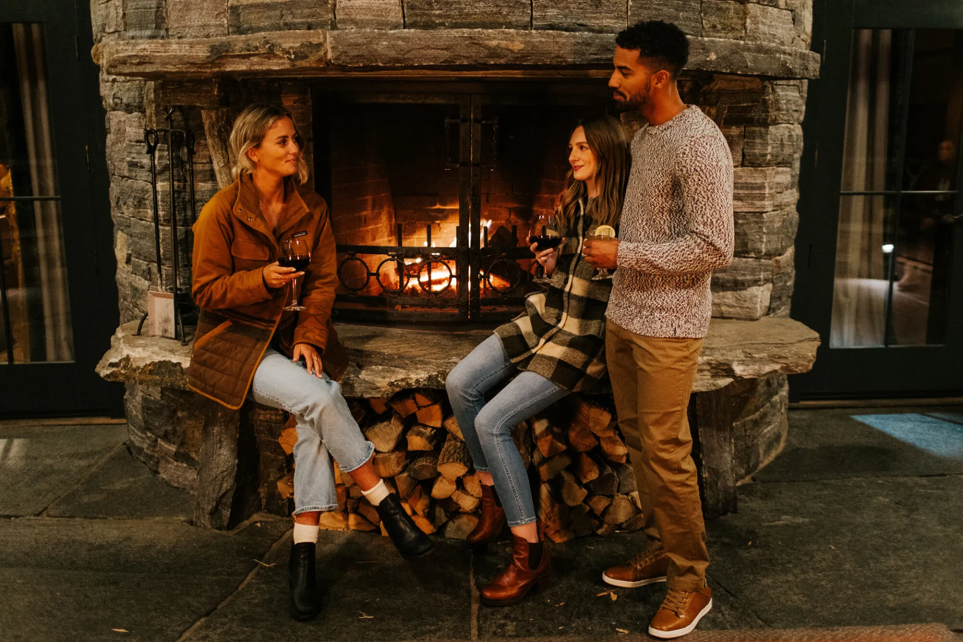 Three friends share drinks at a fireplace. 