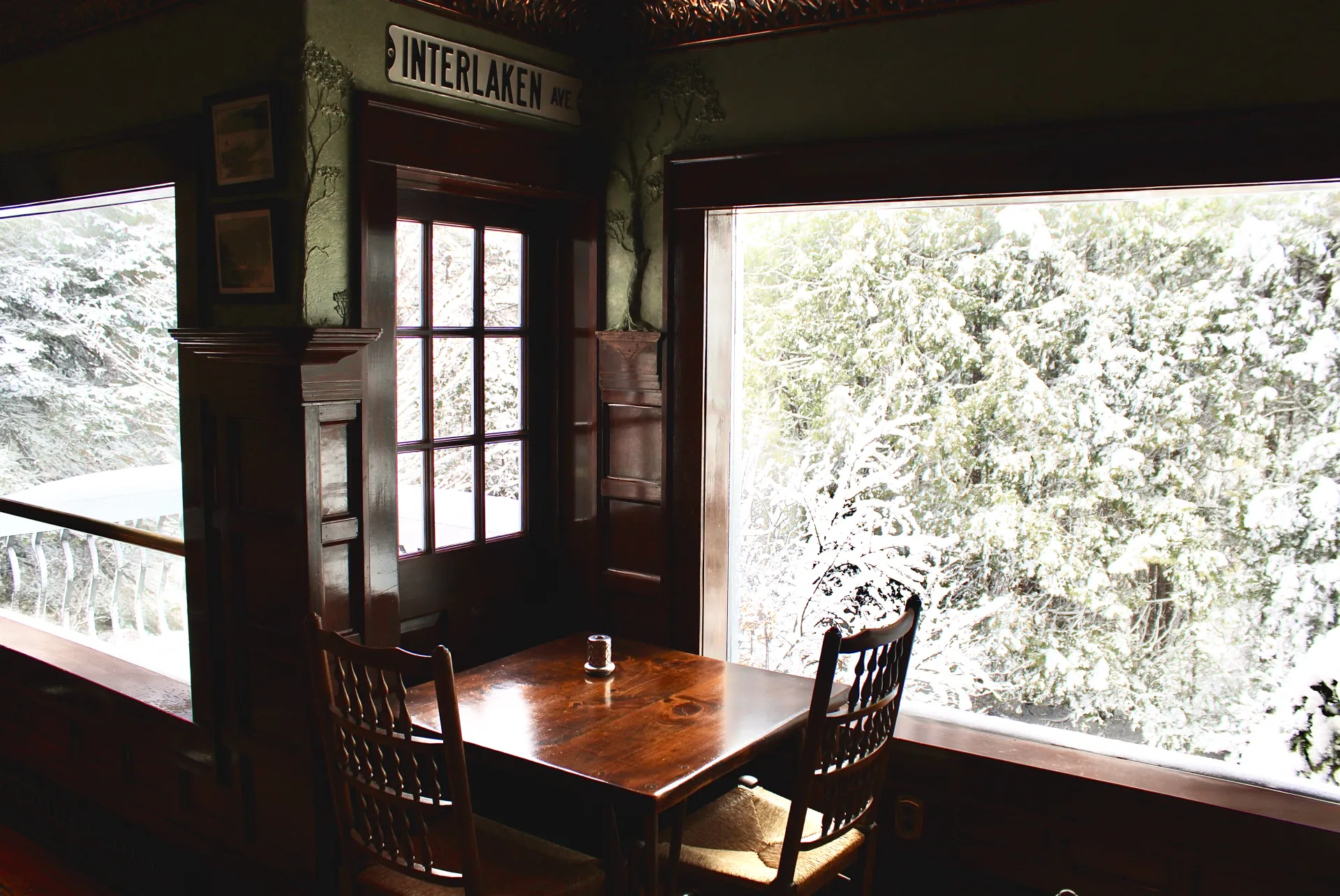 A large window out to the surrounding snow covered trees from the dark dining space.