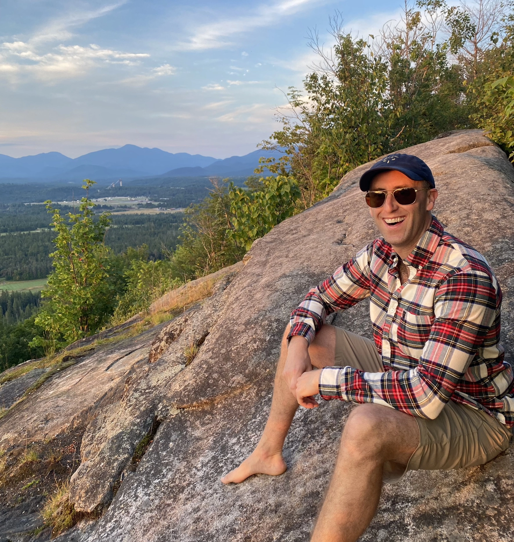 A bare foot man in sunglasses and a flannel sits atop a rocky mountain top.