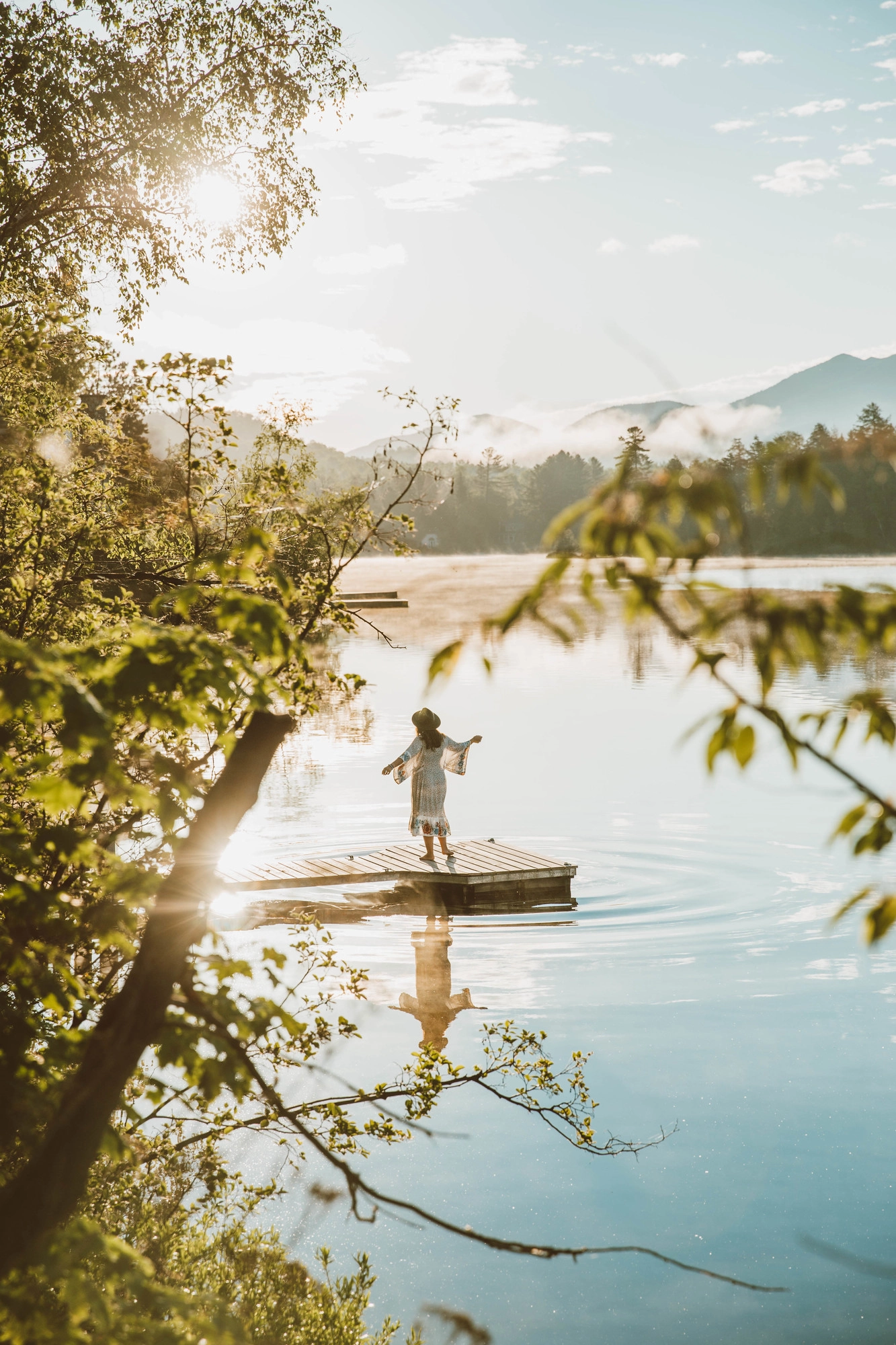 Woman wearing white dress embroidered with flowers and hat seen from between tree branches dancing on lake dock facing mountain views