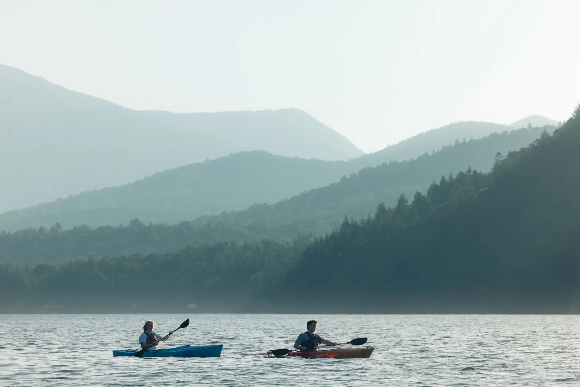 Two kayakers paddle on Lake Placid with layered mountains in misty background