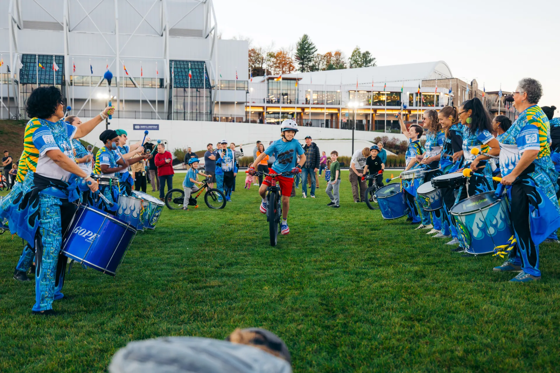 Boy rides bike between rows of a performing drum line as other kids wait their turn and adults look on at Olympic Oval
