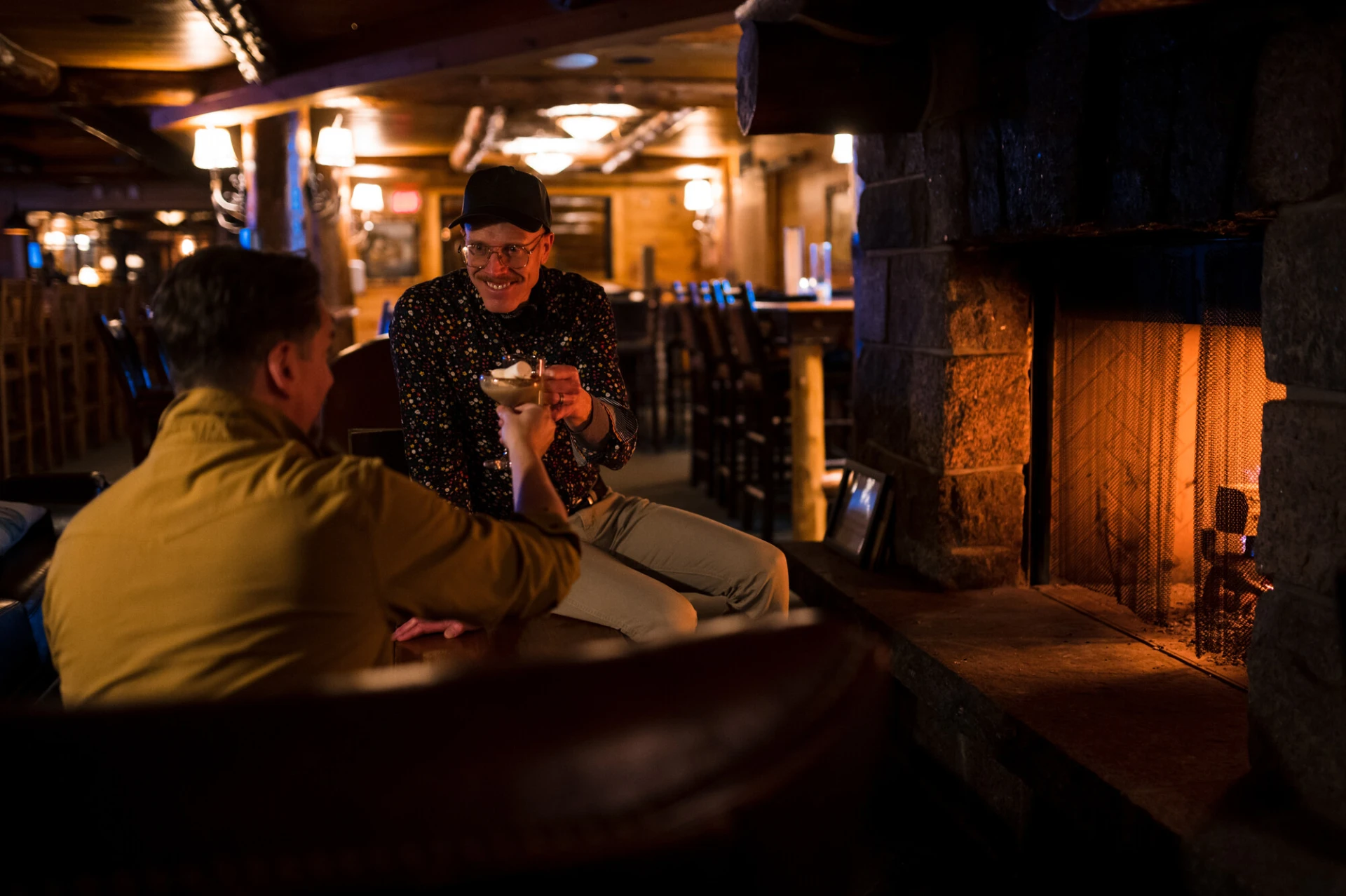 Two men enjoy drinks by a fireplace in a dark bar