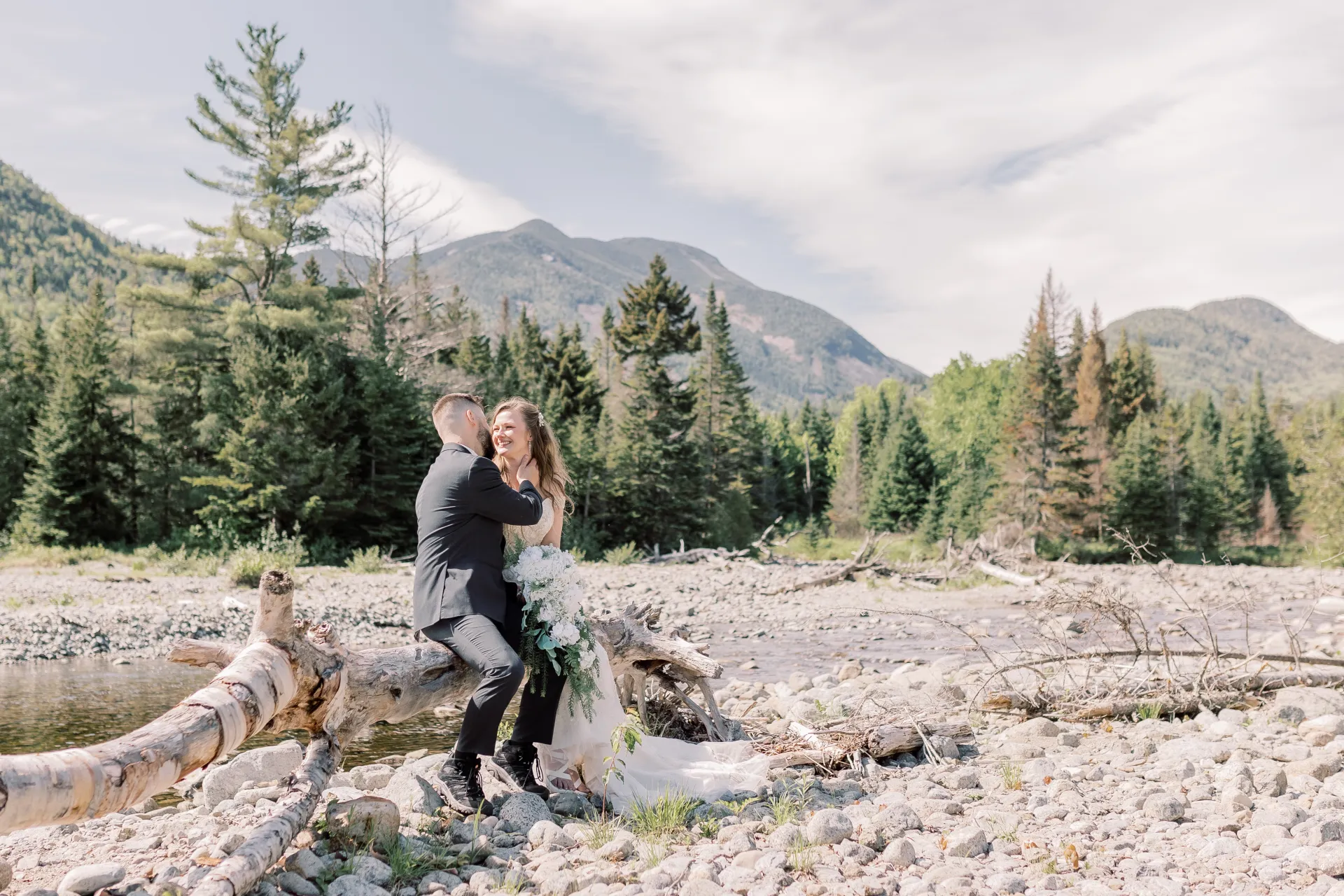 bride and groom snuggle on a fallen tree by a rocky river bed with mountain backdrop