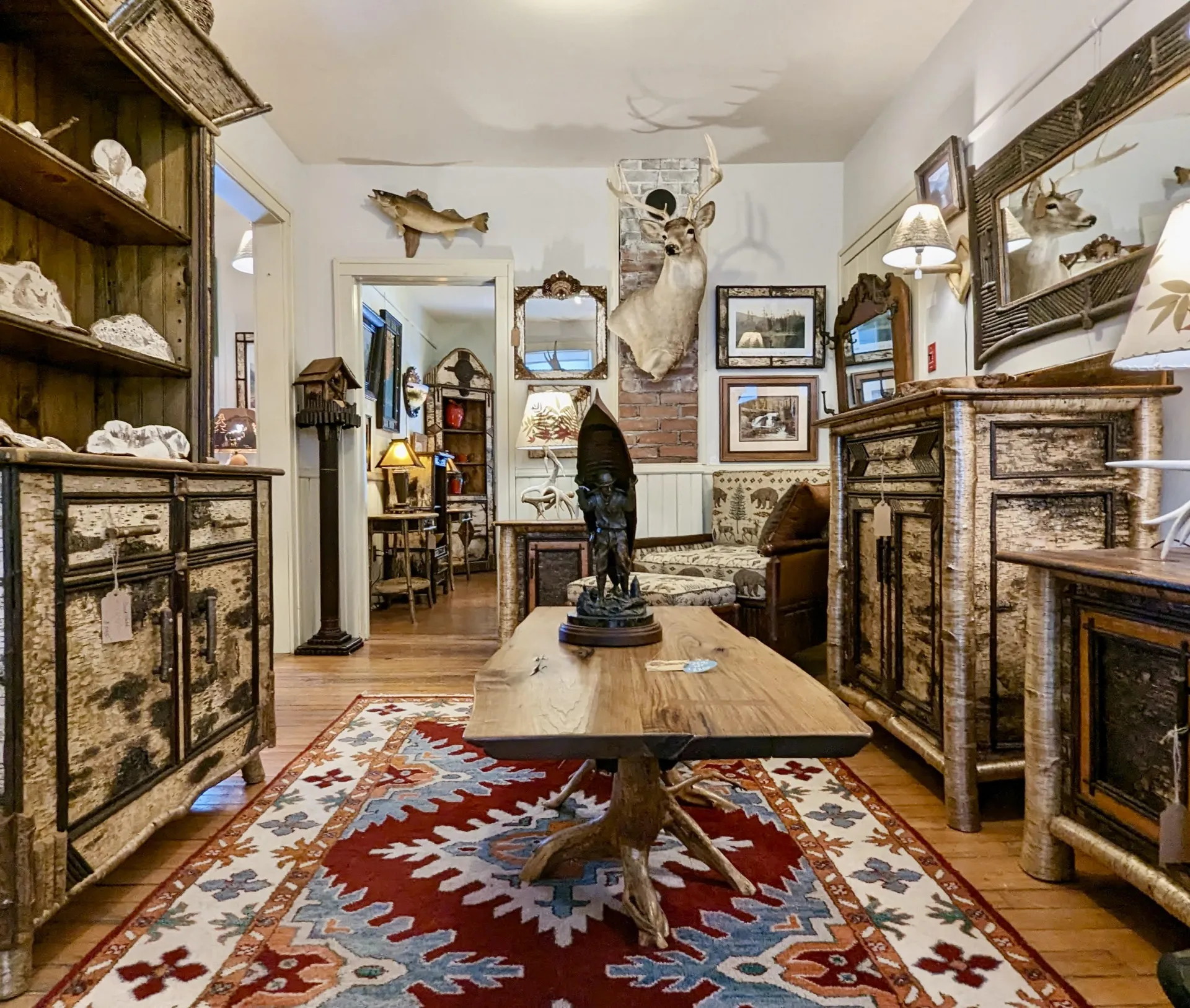 Large hutch and matching dresser made from white birch bark, a coffee table with live edge top and root ball base, a carving of a man carrying a canoe, mounts of various animals hang on the walls punctuated with mirrors surrounded with various barks.