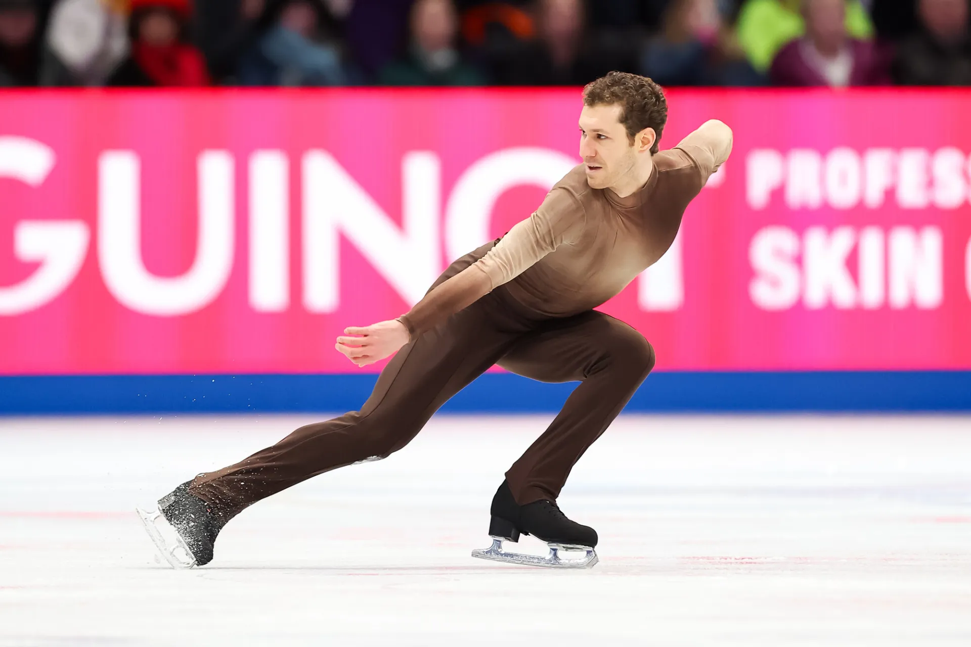Male figure skater in brown long sleeve and long pant spandex suit performs a crouched move on ice