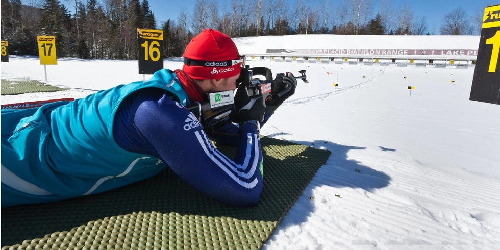 A biathlete takes aim with a rifle while lying down on a pad on snow.