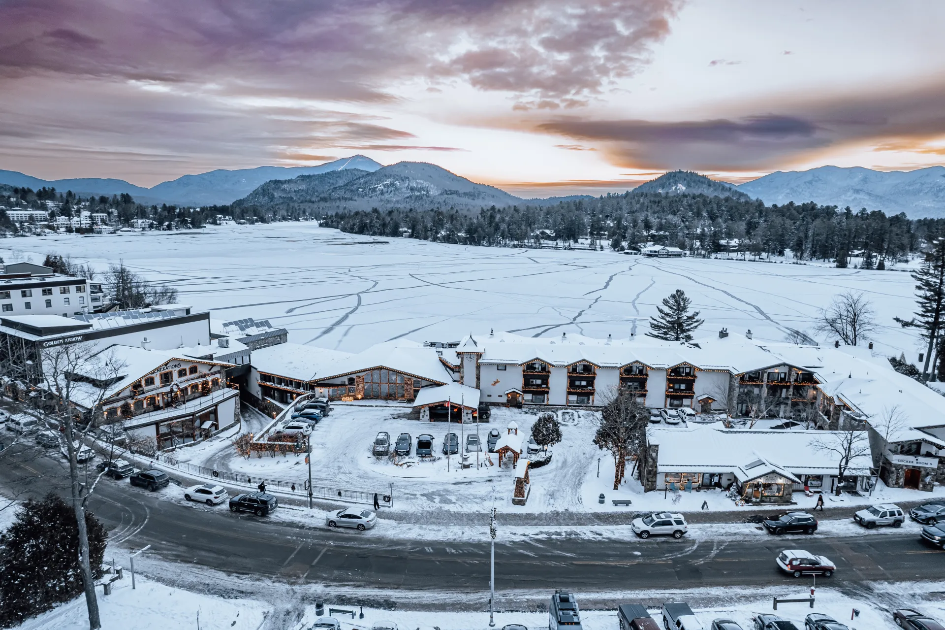 An aerial view of Golden Arrow Lakeside Resort blanketed in snow next to Mirror Lake and the mountains beyond.