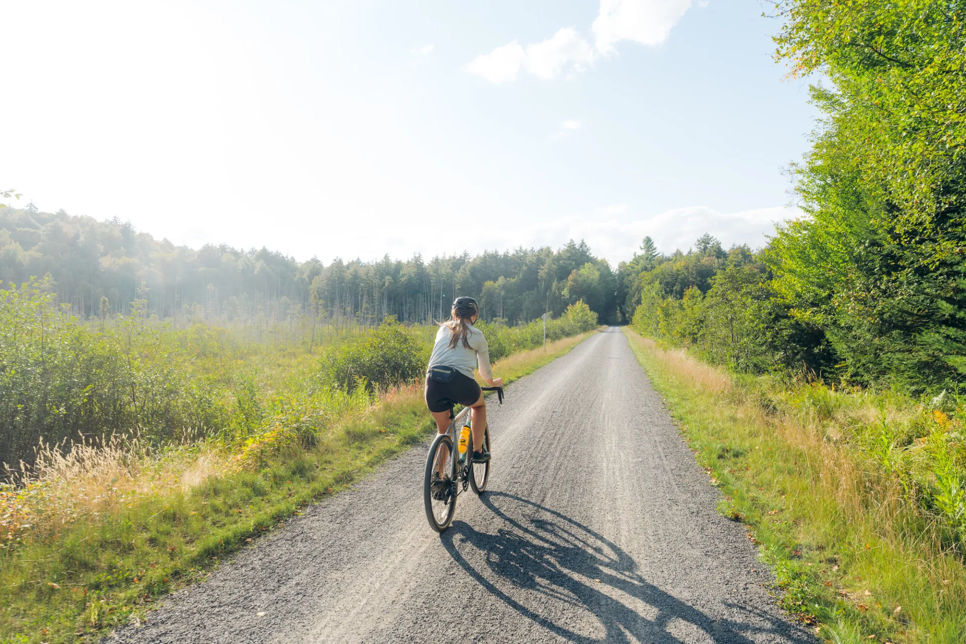 A person rides a bicycle down a long, gravel path through a lush green forest under a bright, hazy sky.