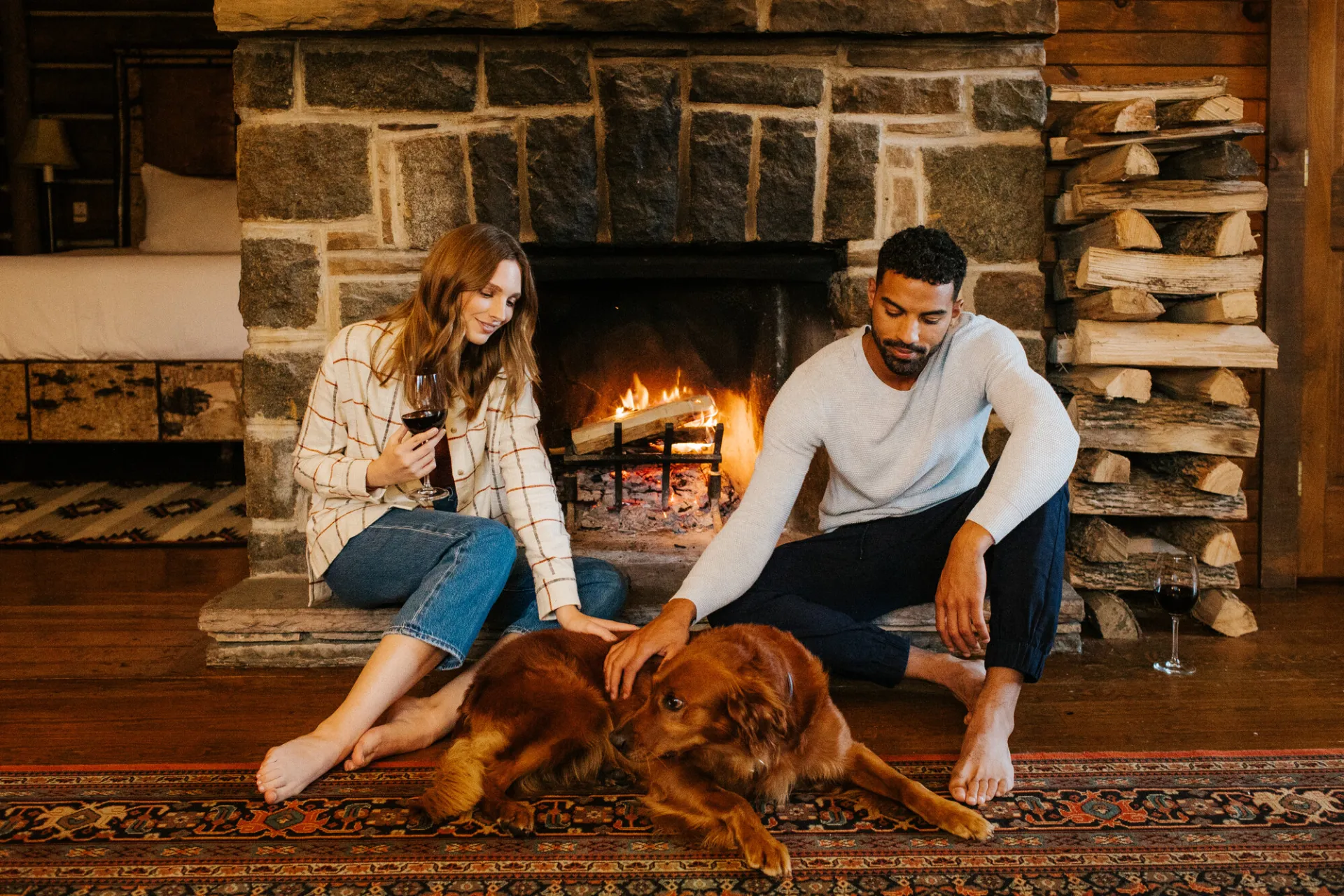 Man and woman sit on hearth of stone fireplace with red dog between them. The woman is holding a glass of red wine