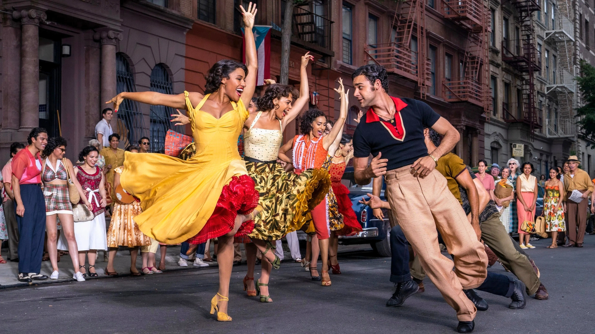 A group of dancers perform a routine on a city street lined, led by a woman in a yellow dress and a man in a polo shirt.