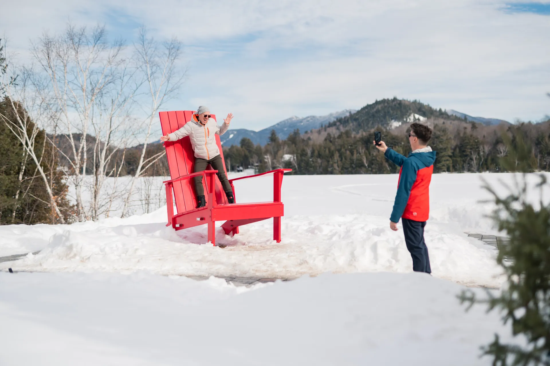 A man takes a photo of another man posing on a giant Adirondack chair in a snowy park with mountains in the background.