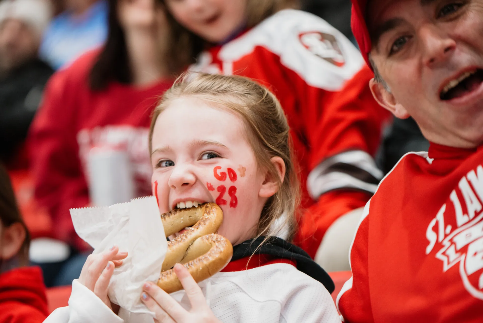 A girl with blonde hair and "Go 28" painted in red on her cheek looks at the camera as she bites into a large soft pretzel.