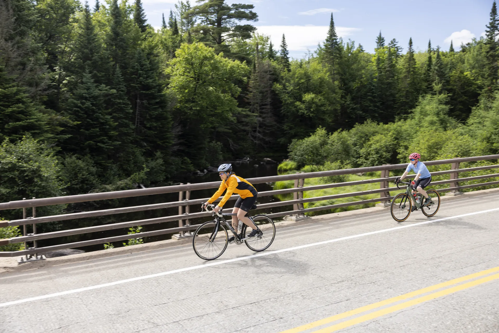 Two cyclists ride on paved bridge with metal railing over river with mix of green trees in background