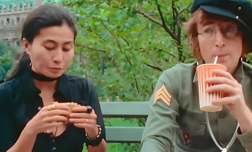 A woman with dark hair eats a sandwich while John Lennon, wearing a newsboy cap and glasses, drinks from a paper cup.  They are sitting on a park bench outdoors.