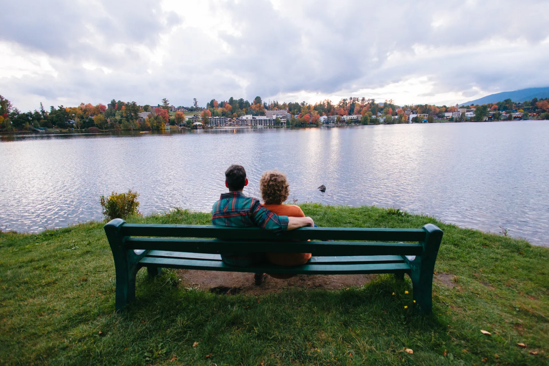 Couple sits on green bench next to lake looking over water at Village of Lake Placid during fall