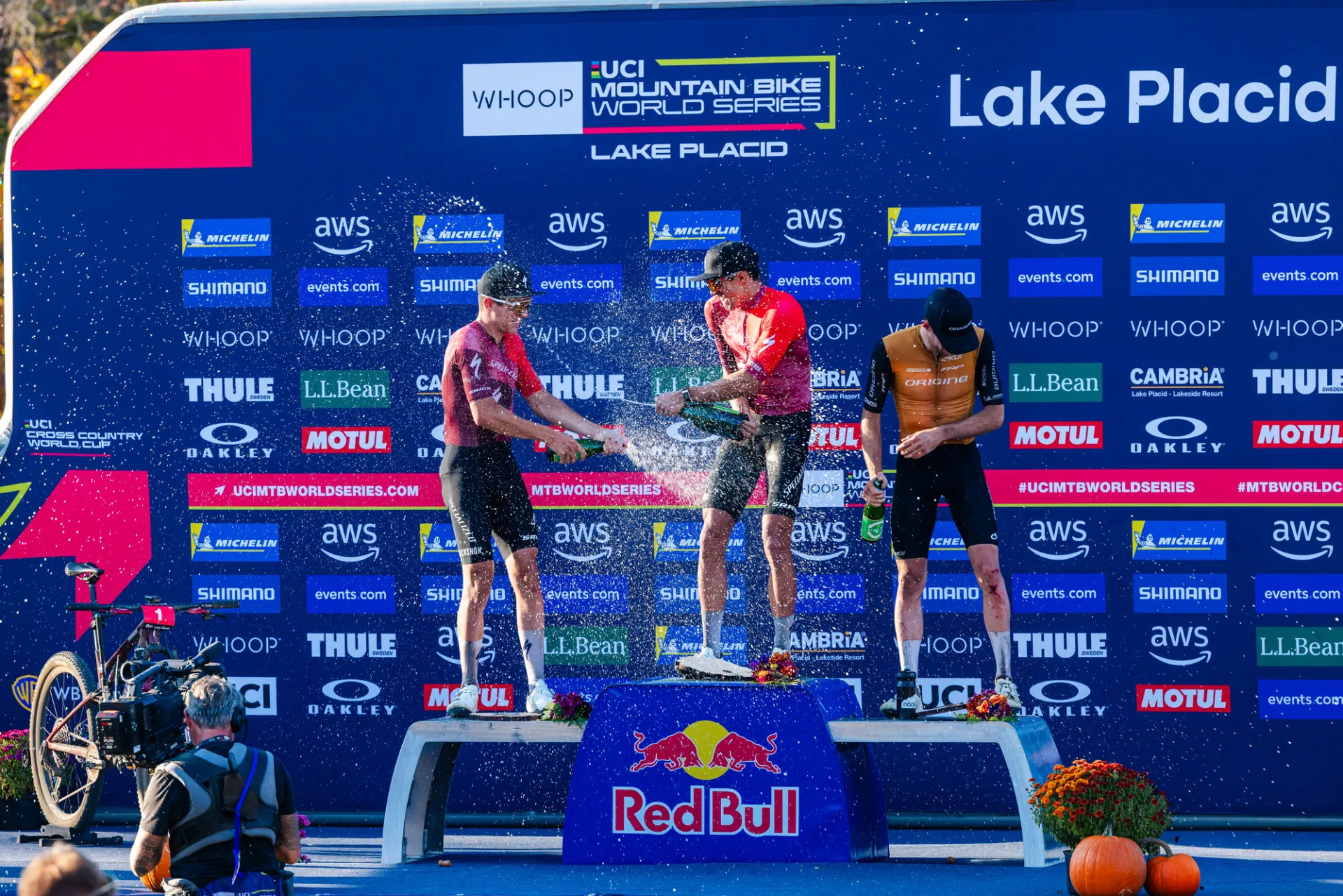 Three mountain bike racers stand atop three stage podium spraying each other with event logos on blue backdrop