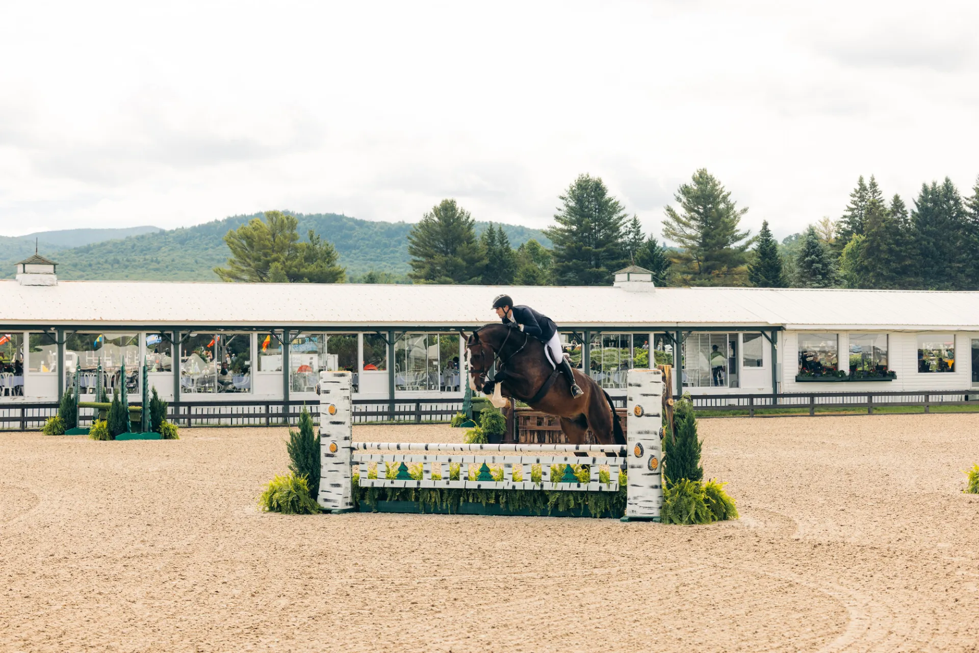 Equestrian rider jumps horse over jump on dirt ring with pavilion building in background