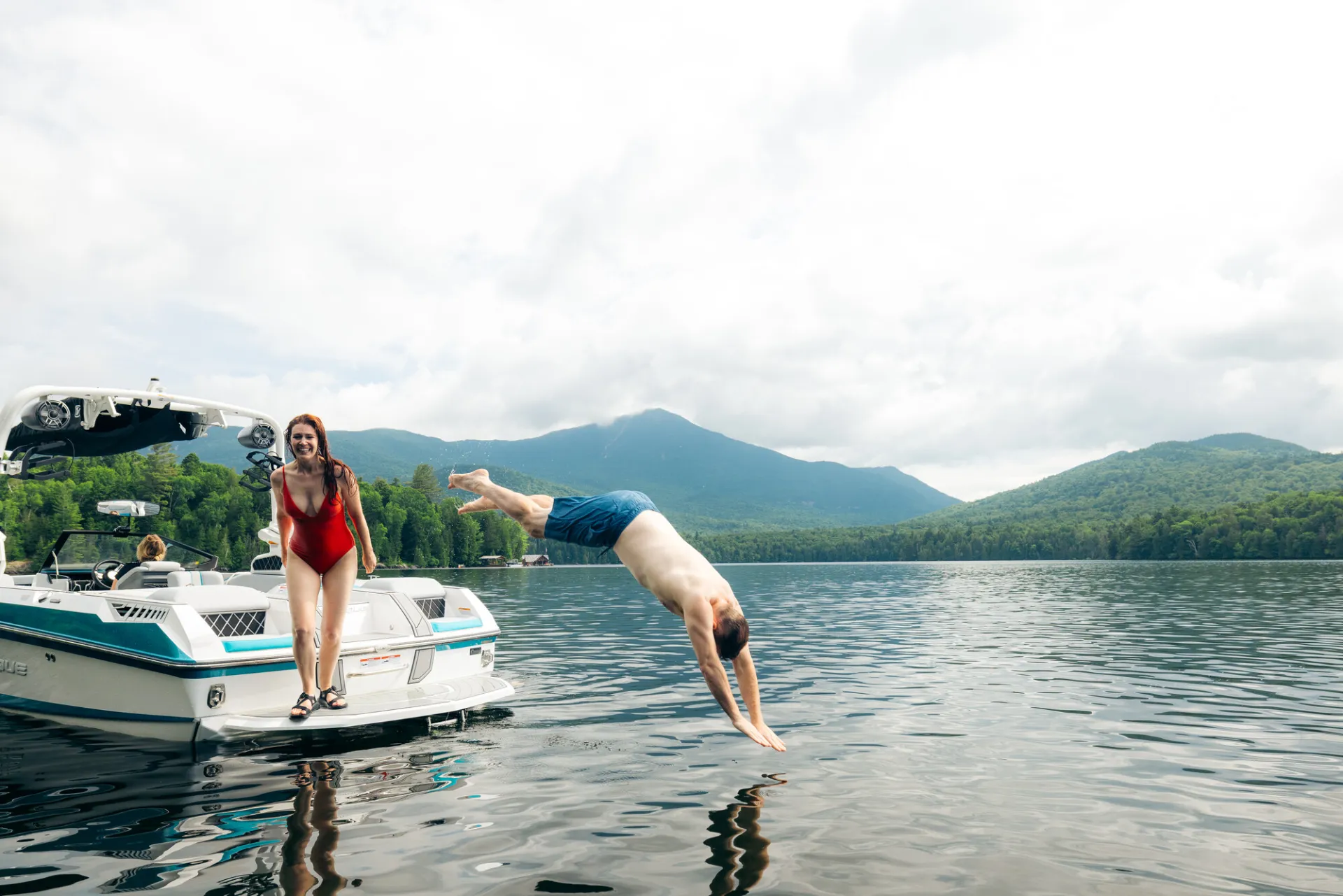Man dives off motor boat into Lake Placid while woman in red bathing suit looks on laughing with mountains in background