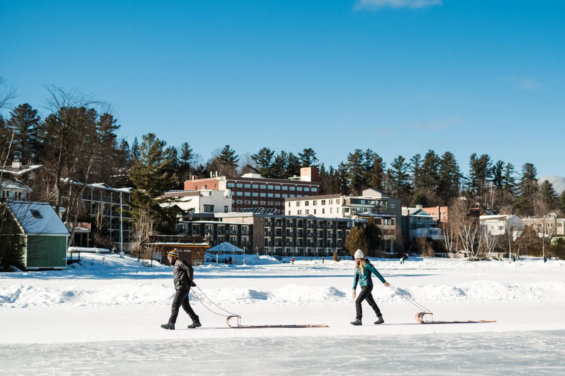 Couple walks across icey lake pulling toboggans with town in the background