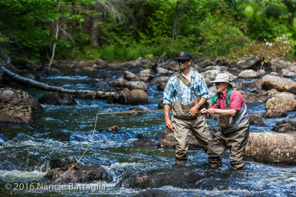 Two men wearing waders stand in a rocky, forested river while one demonstrates fly fishing techniques with a rod and line.