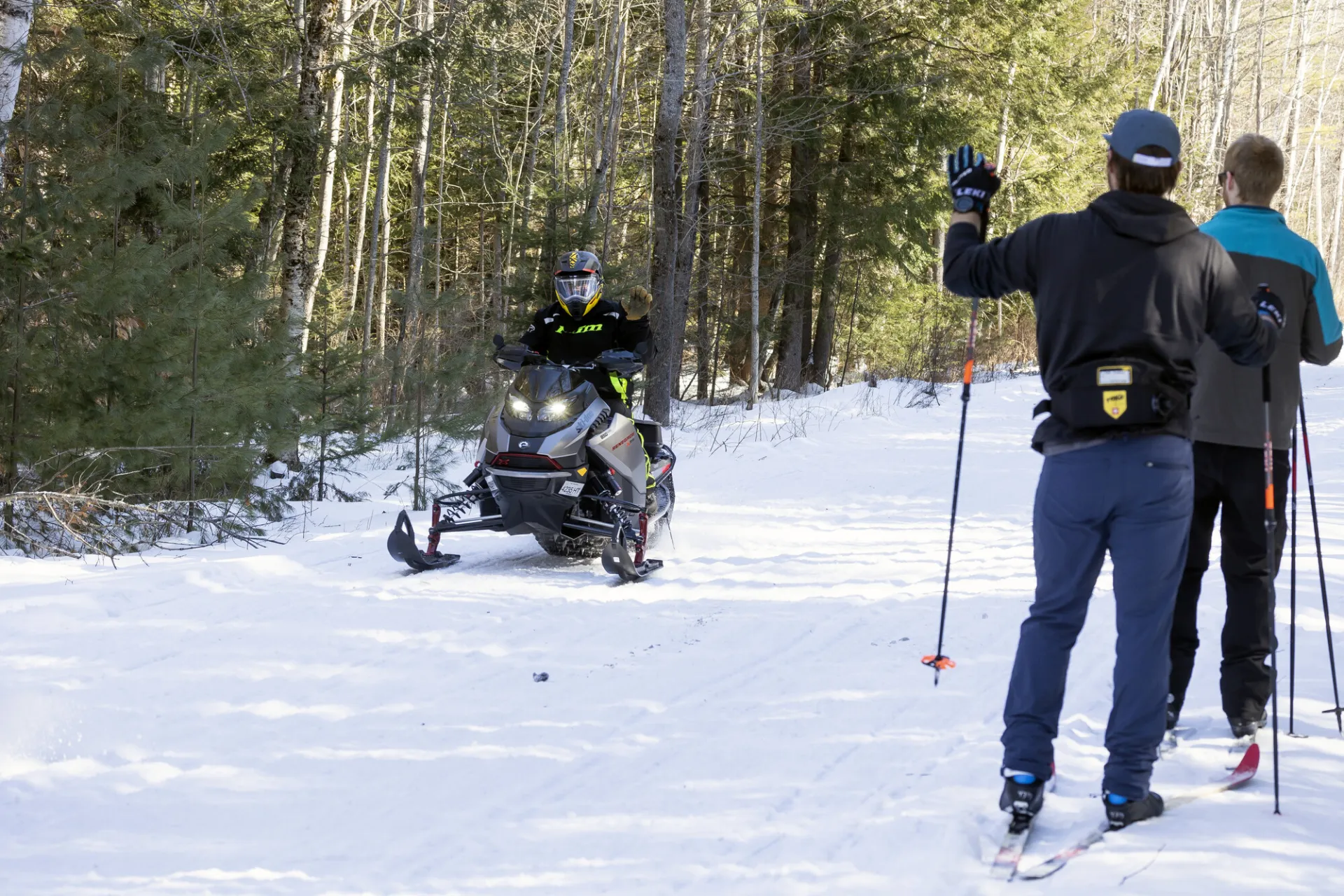 Skiers wave to a snowmobiler on a wide snowy pathway. 
