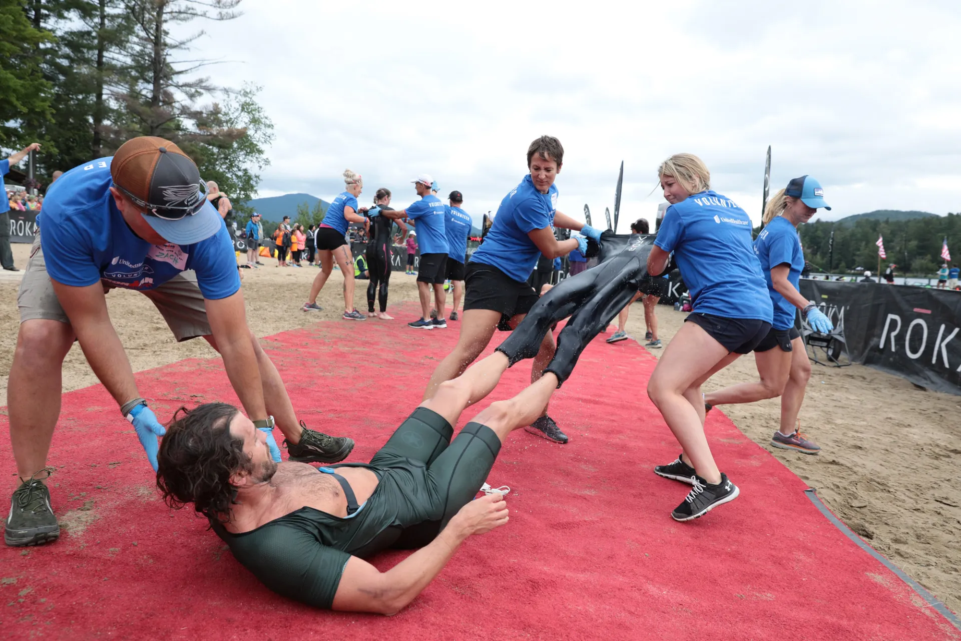A swimmer lies on a red carpet on the ground while three volunteers in blue Ironman t-shirts remove his wetsuit, one holding his shoulder and two pulling the suit off his legs