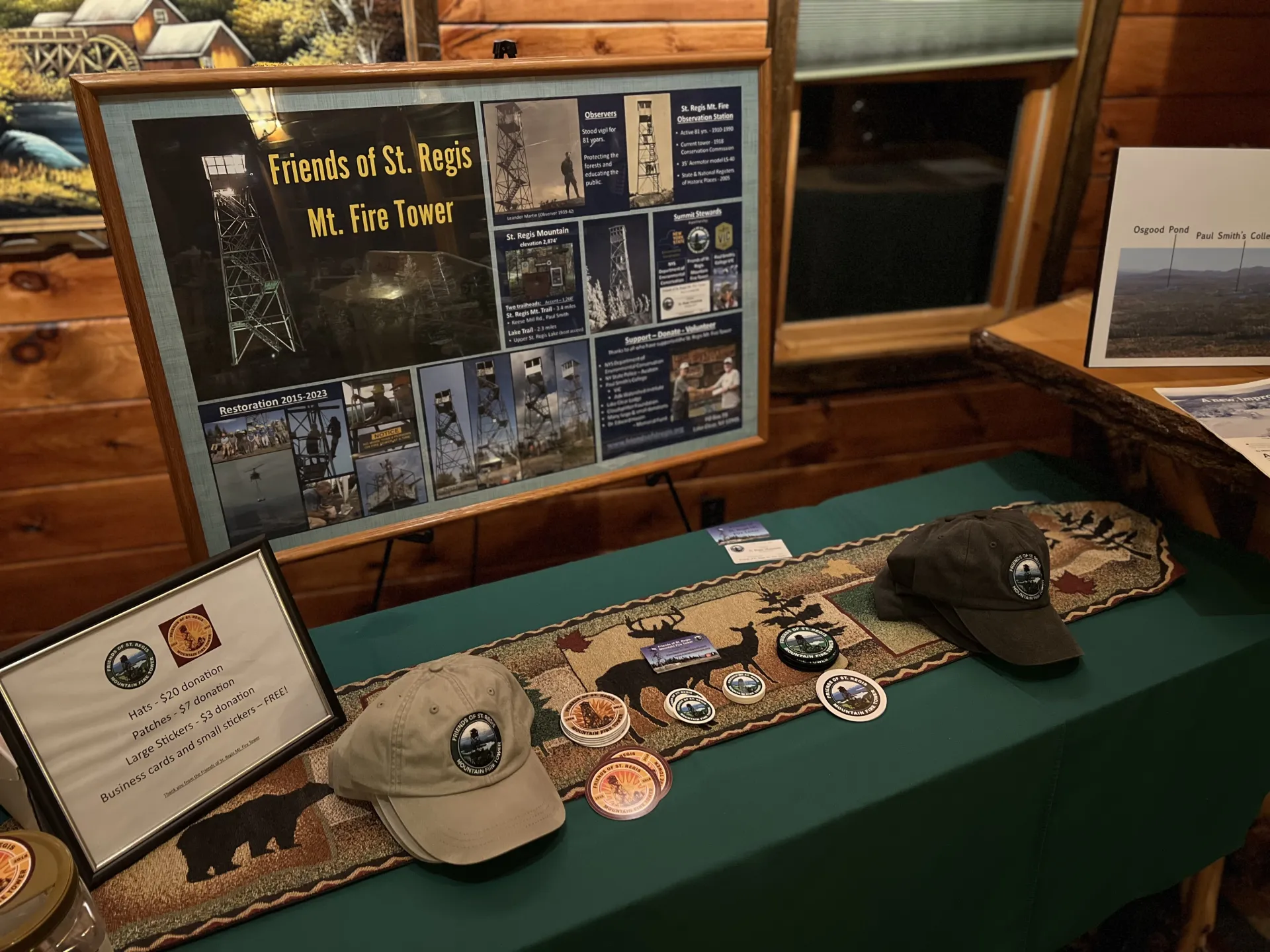 Display table with information about the restoration of the St. Regis Mountain fire tower, including photos and details about its history and construction.  Also shown are hats, patches, and stickers for sale to support the restoration.