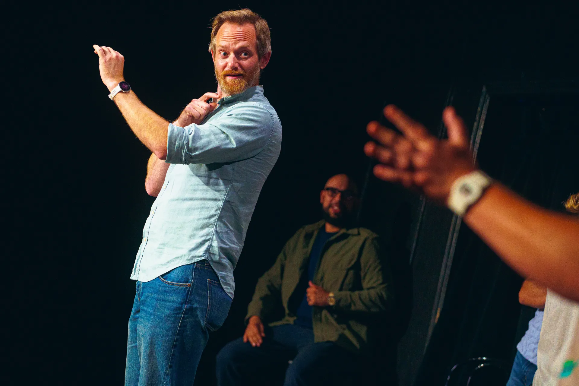 Bearded man in dress shirt and jeans holds up one hand and looks and points in the direction of someone out of image adjacent