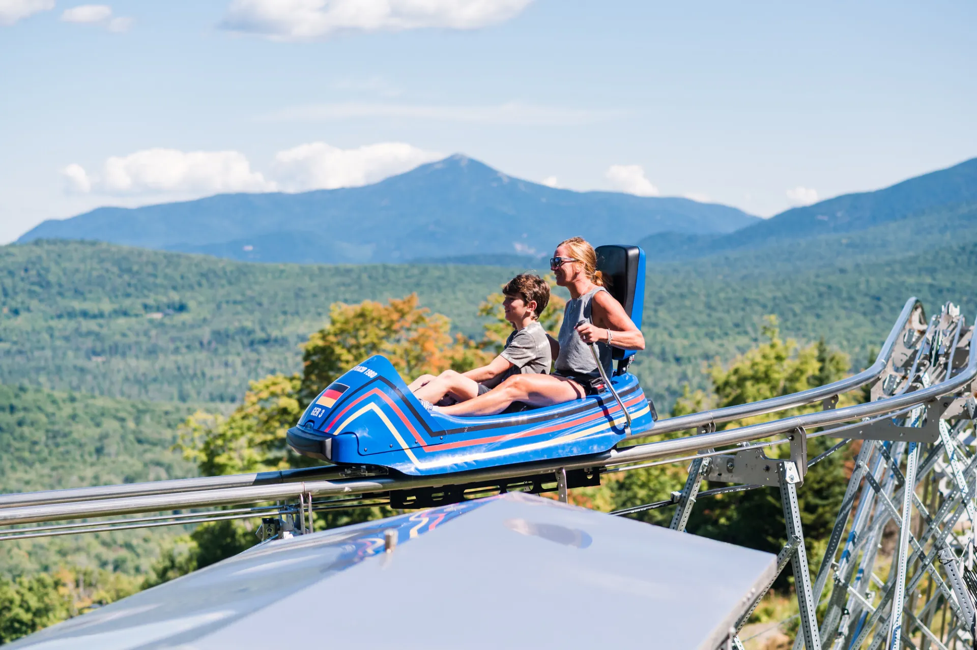 A woman and a boy ride a blue, bobsled-style coaster on rails, with mountain views beyond.