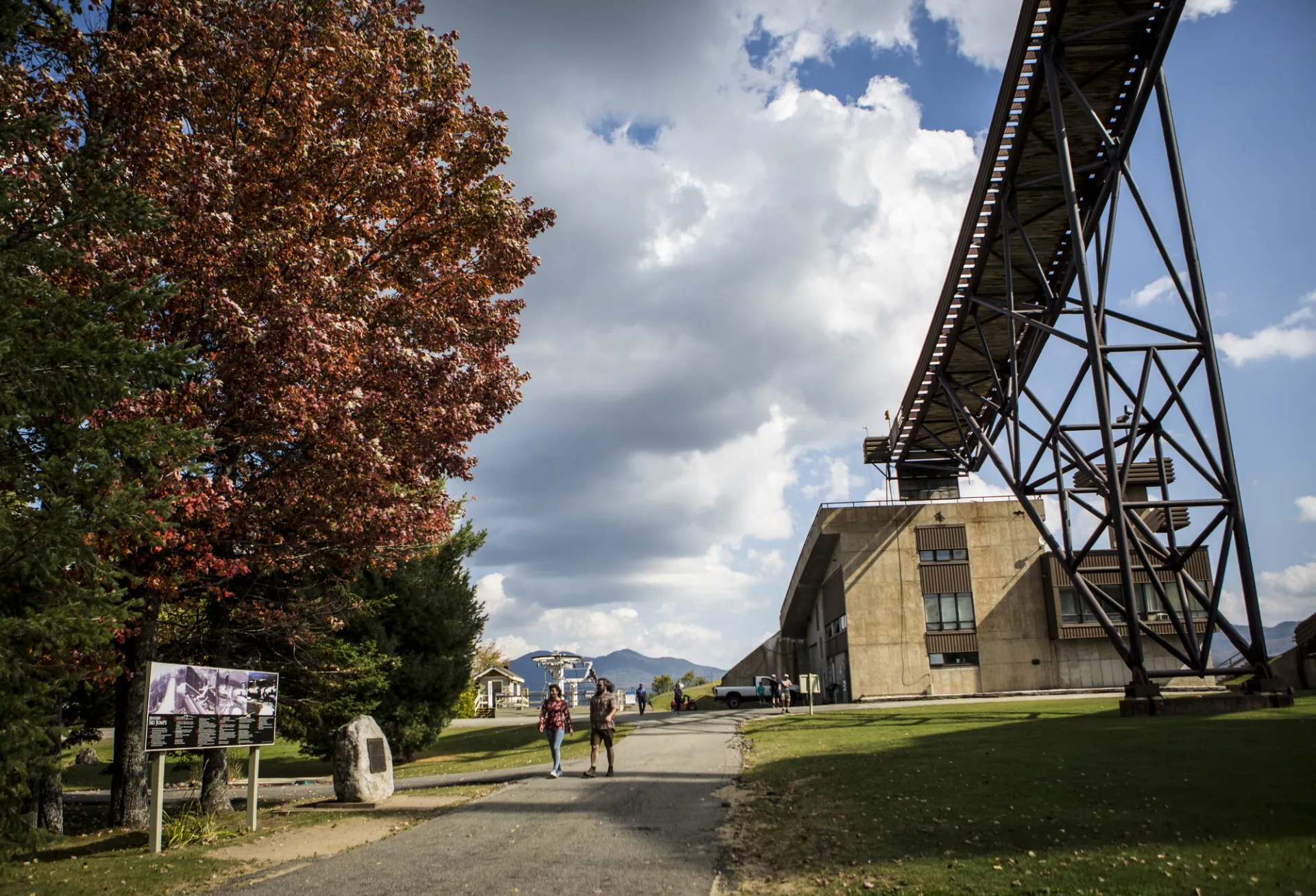 A couple walks on a path near a tree with red leaves and the towering Olympic ski jump.