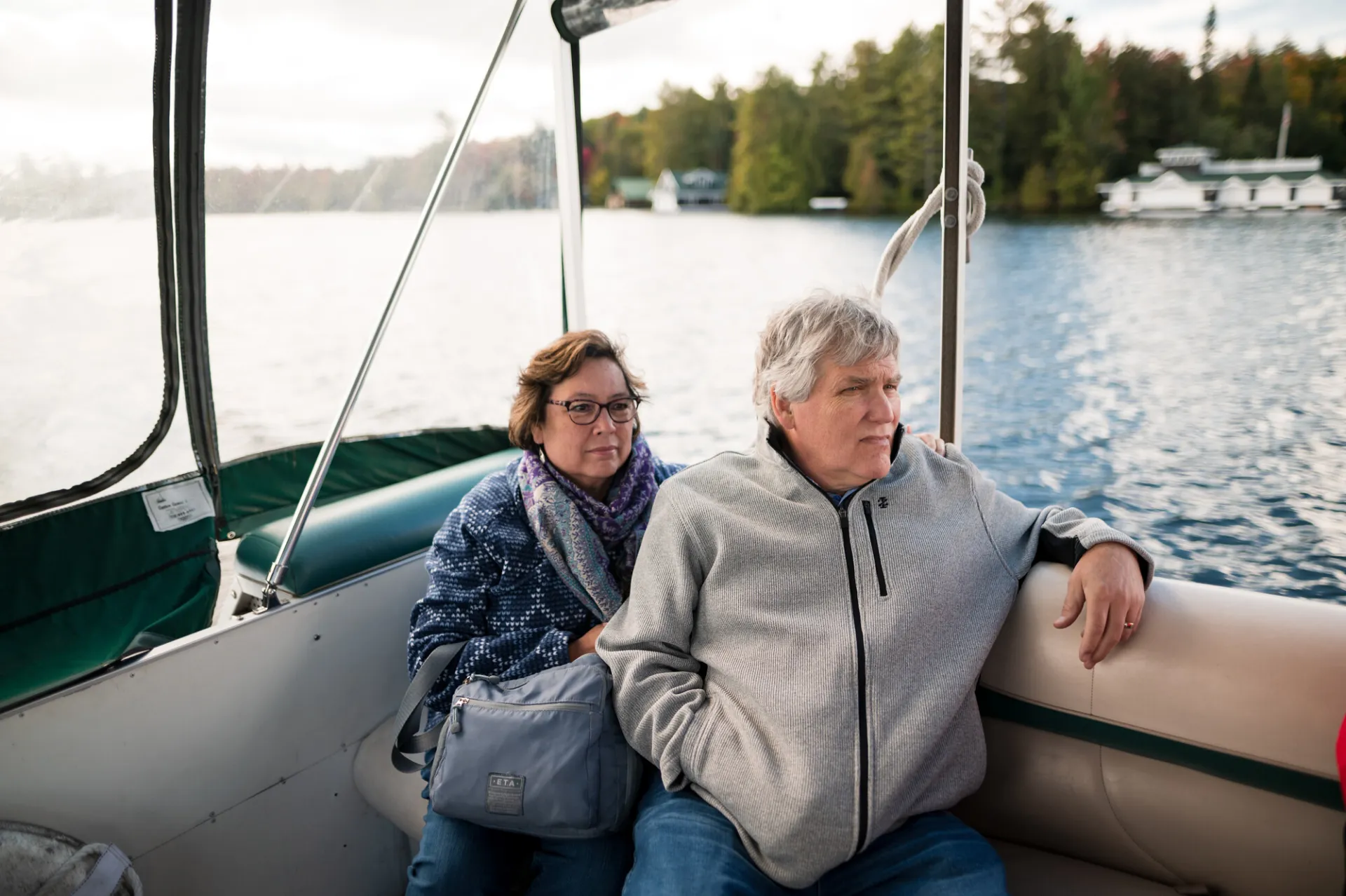 A man and woman in jackets sit on a boat on a lake during fall.