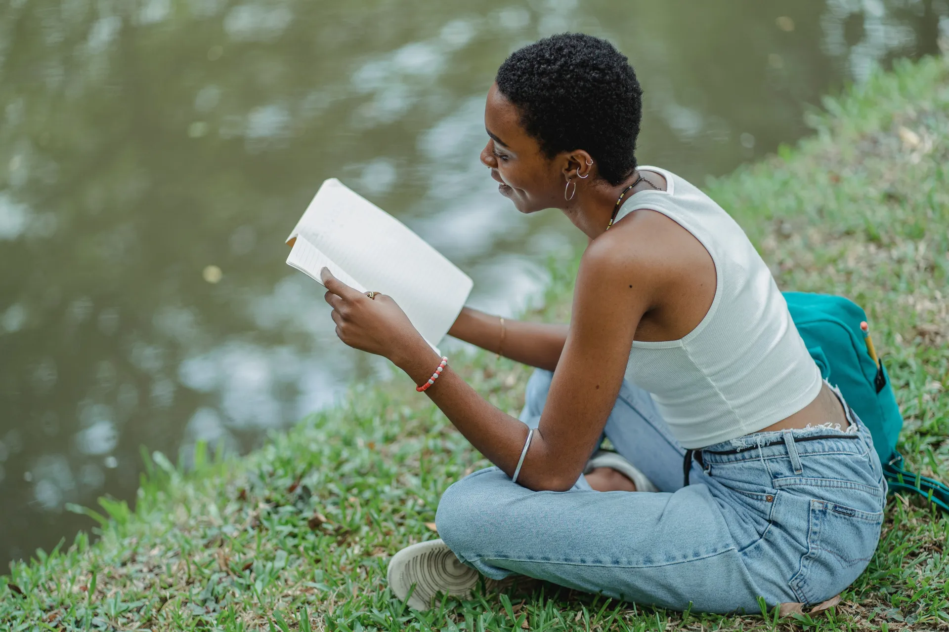 Young woman sits next to lake on grass reading a book wearing jeans and white tank top