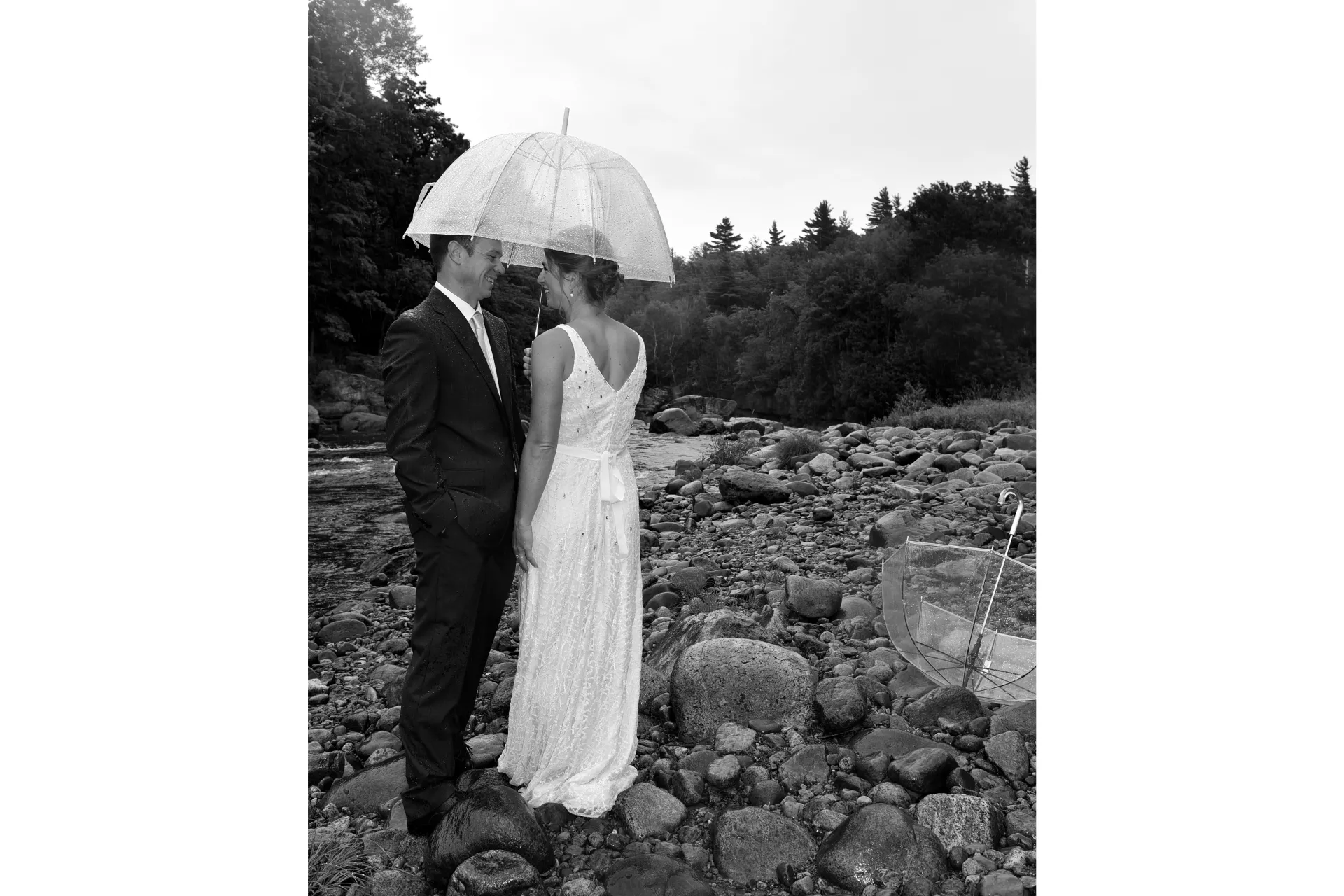 A bride and groom stand by a riverbed under a clear umbrella in black and white.