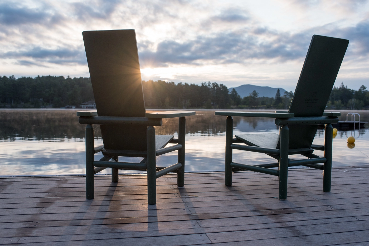 Tall backed chairs on a dock over looking a smooth lake with mountains in the distance.