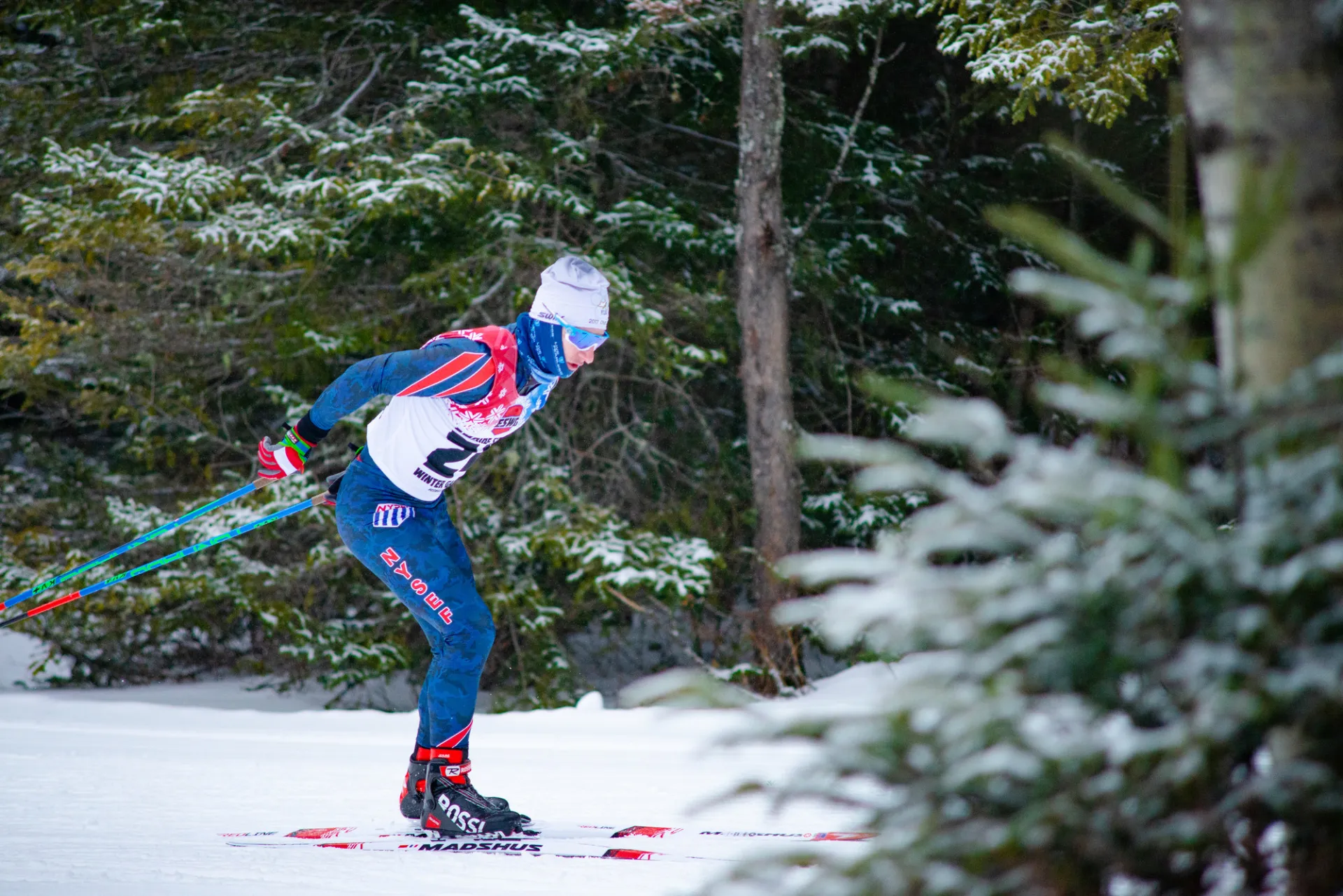 A cross-country skier in a blue and red racing suit and a white beanie glides along a snow-covered trail through a pine forest.