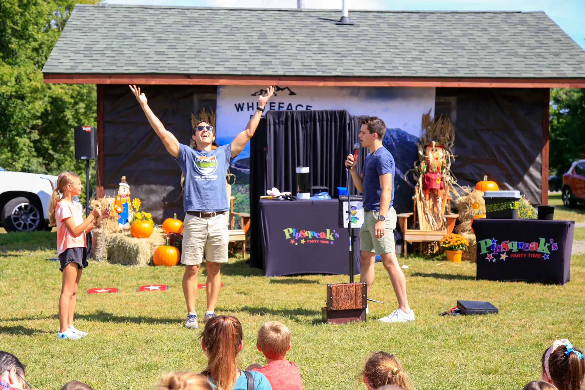 Two men stand in front of crowd of children sitting on grass at fall event.