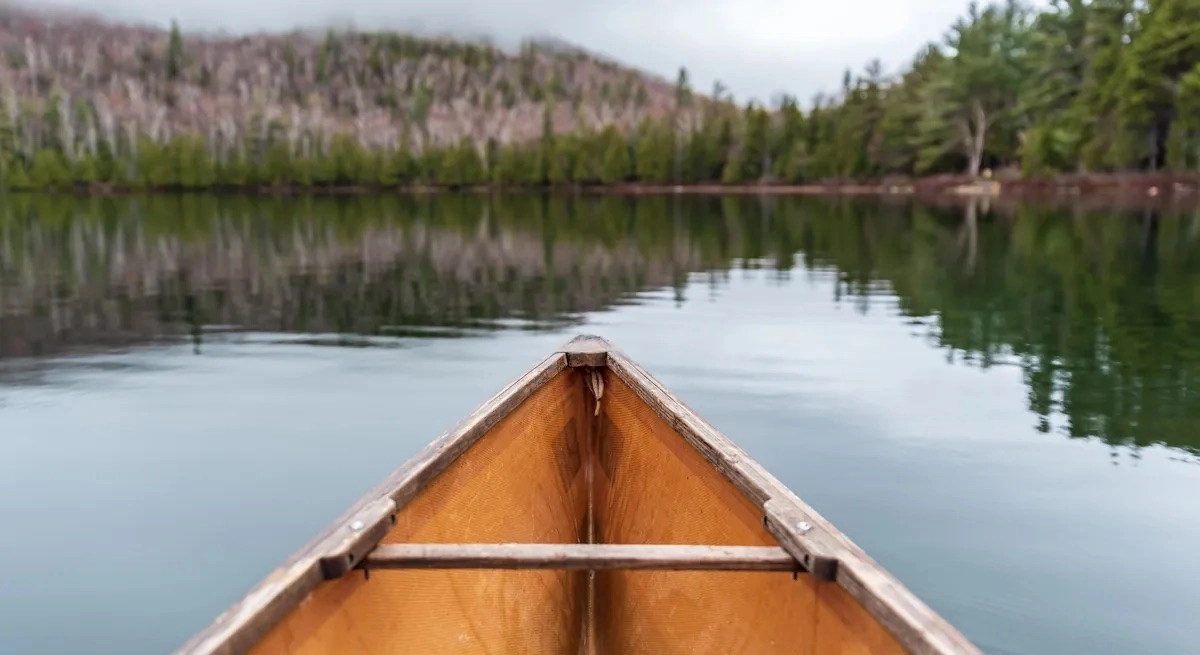 View from the front of a canoe on a calm lake, surrounded by trees and a distant mountain.