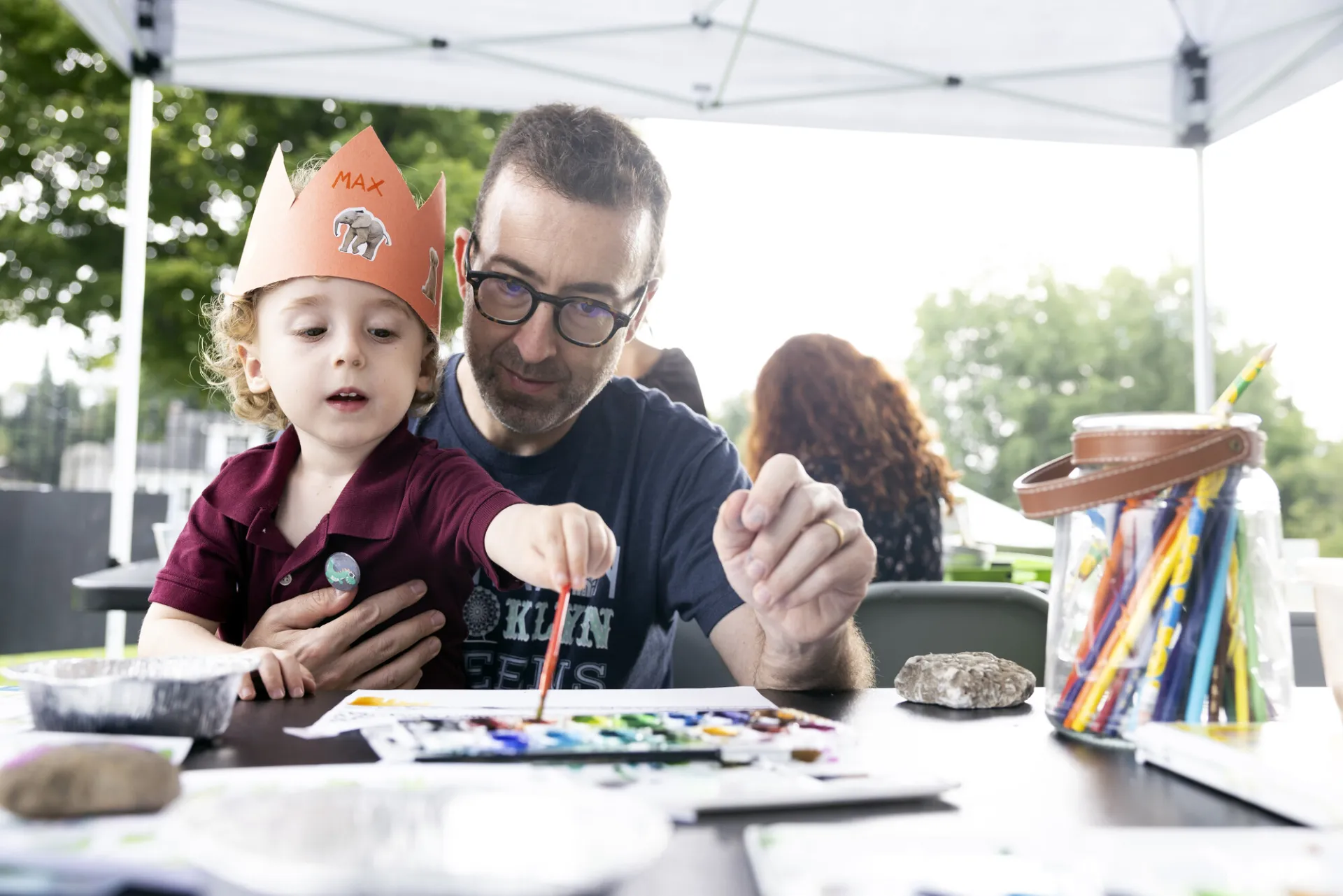 Little boy wearing paper crown sits on man's lap dipping paintbrush in watercolors