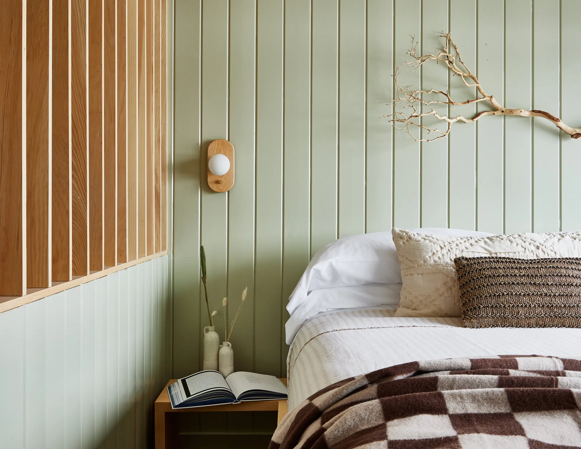 Mint green walls with feature slatted wall and cozy bedding.  A book lays open on the night stand under a lamp.