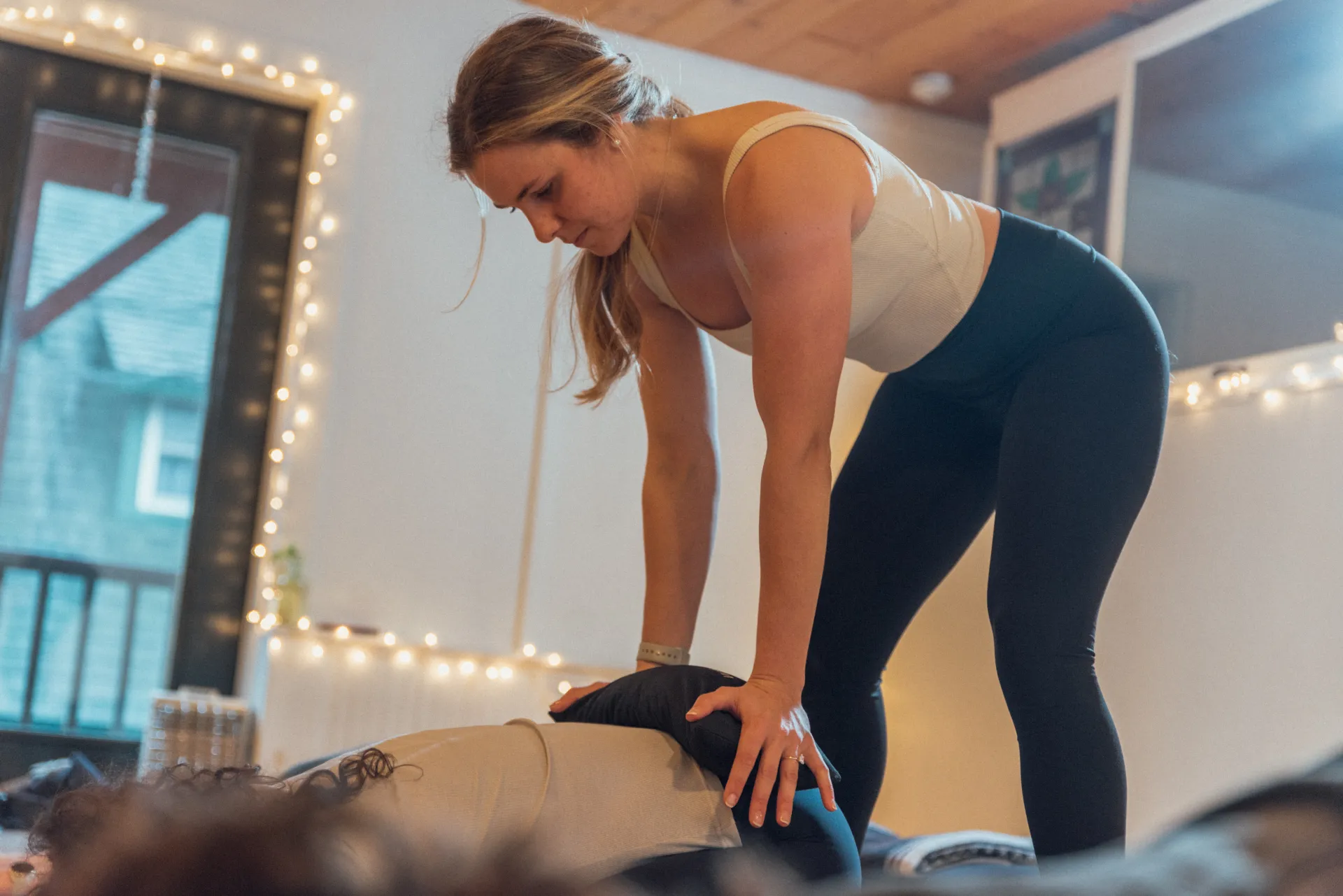 Yoga instructor in white tank top and black yoga pants presses down on the lower back of a person on the floor bent forward in a child's pose with a window and twinkle lights in the background
