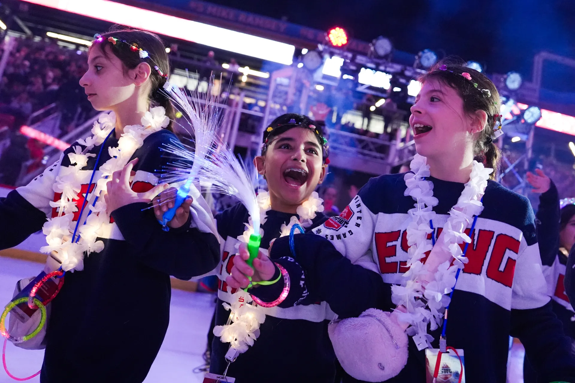 Three smiling children wearing "ESWG" sweaters celebrate with light-up wands during an opening ceremony.