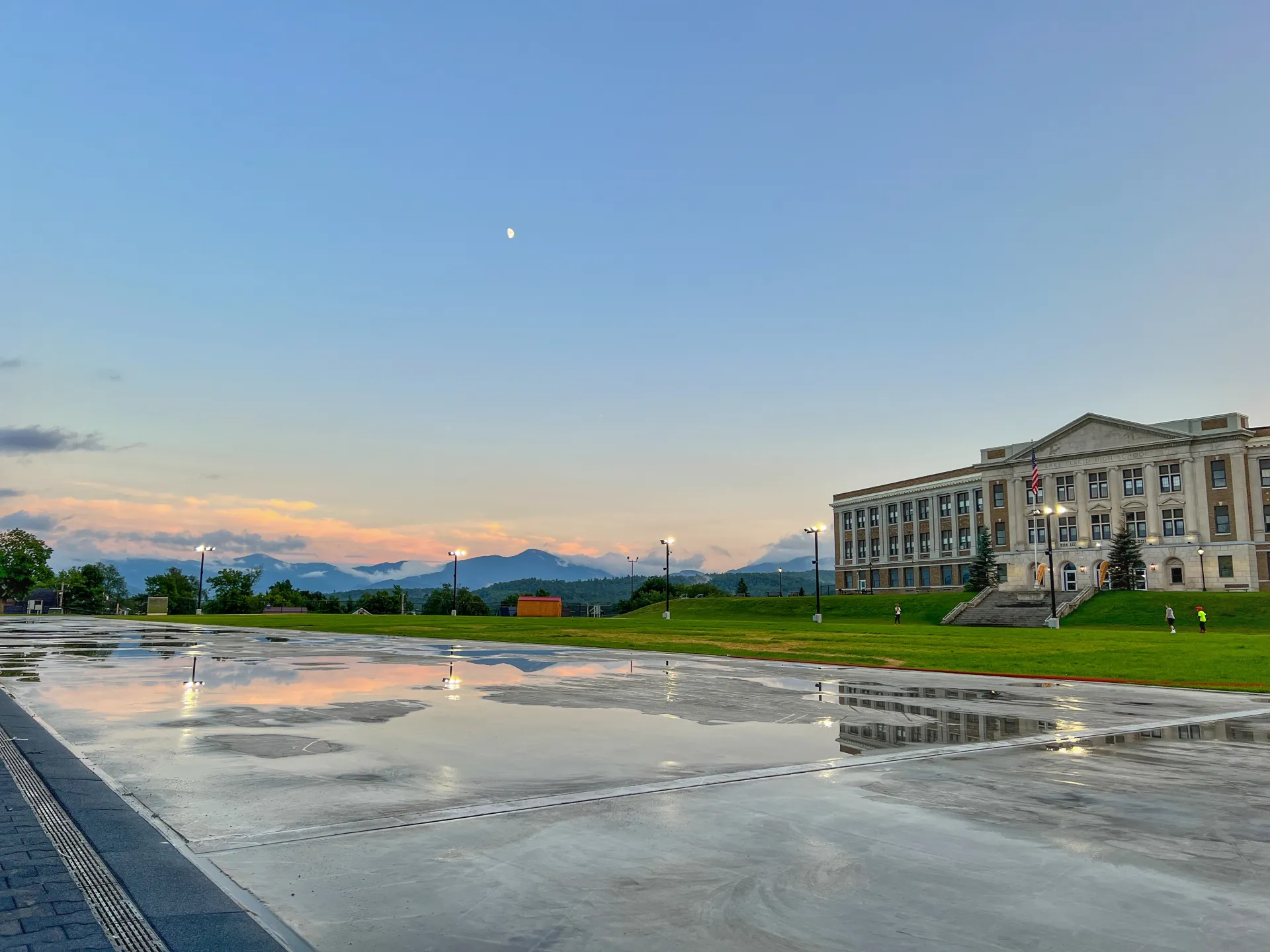 Puddles of rain glisten on a paved outdoor skating track. The moon gleams in the dim blue sky above.
