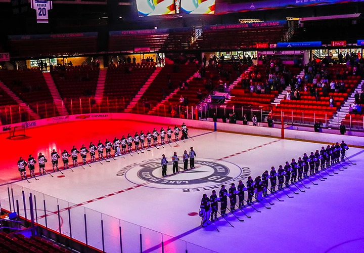 Two teams of female hockey players lined up opposite one another on the ice with officials in the middle under colorful light
