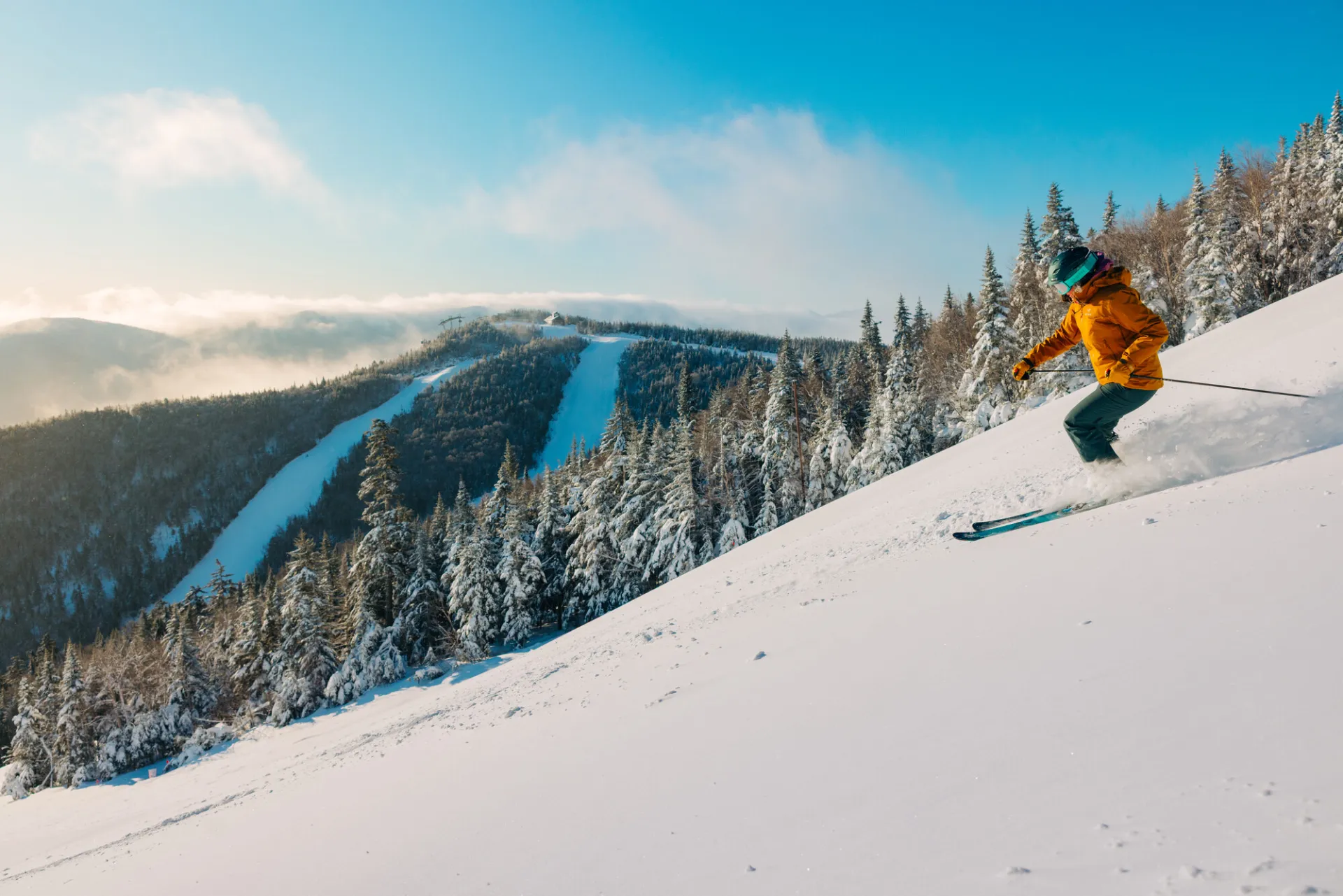 Person in orange ski coat and ski gear descends snowy slope on largely blue sky day with additional mountain peak with snowy runs in bakcground