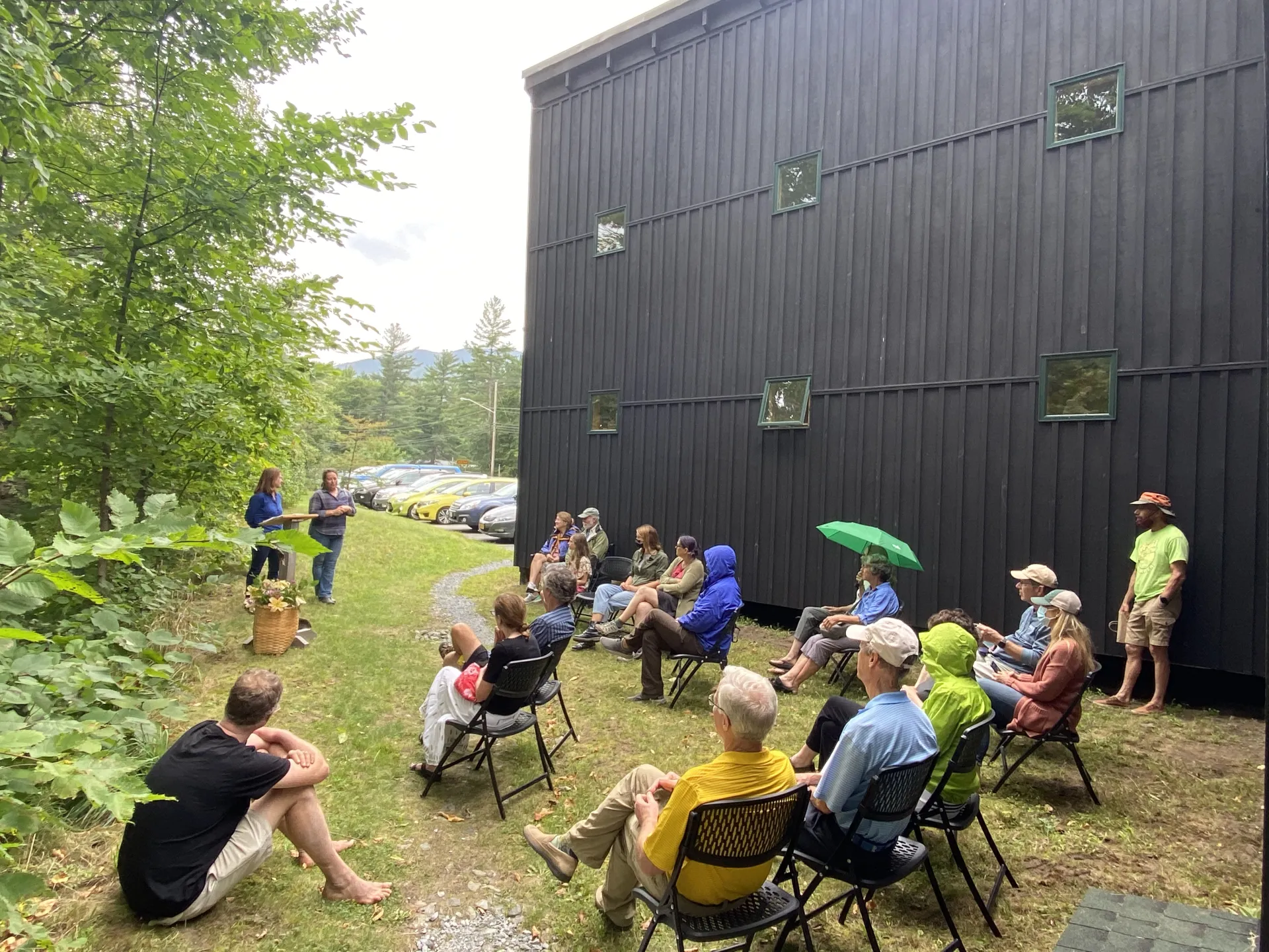 A group of people are seated in chairs outdoors, listening to two people giving a presentation.  A dark building is in the background.