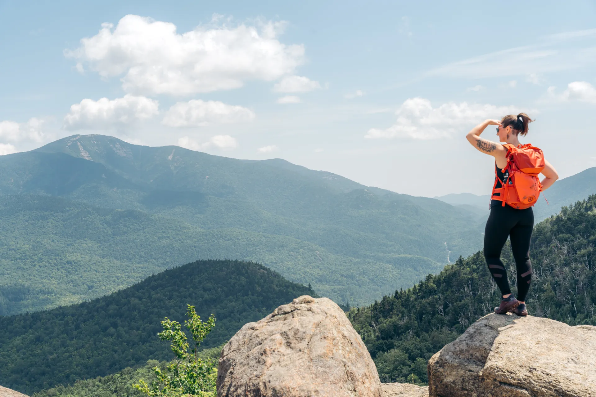 A woman standing at the summit of a hike, looking out at the mountain views.