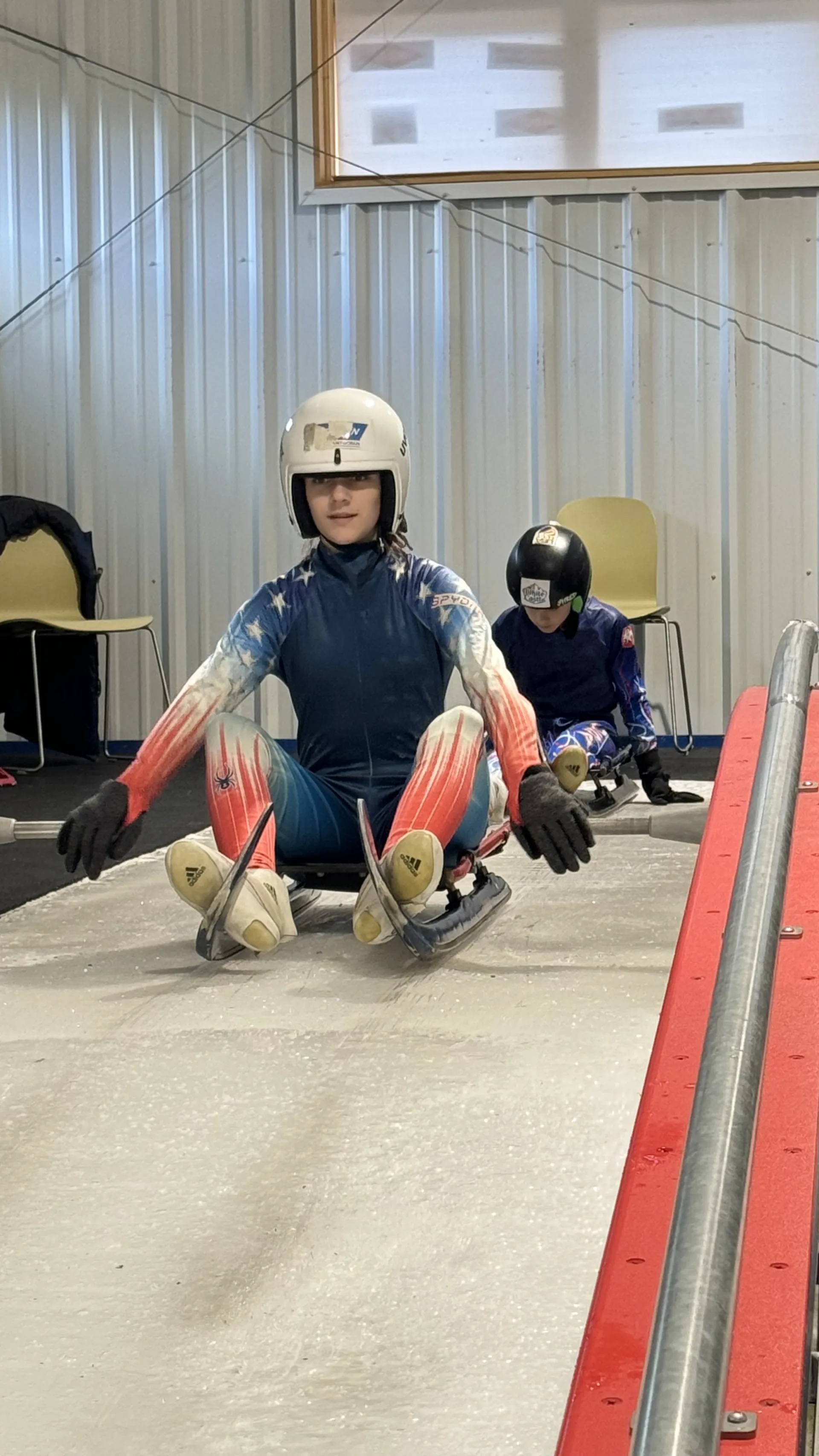 A young woman in a USA luge uniform sits on a luge sled on an indoor track.  Another young person is visible behind her, also on a sled.