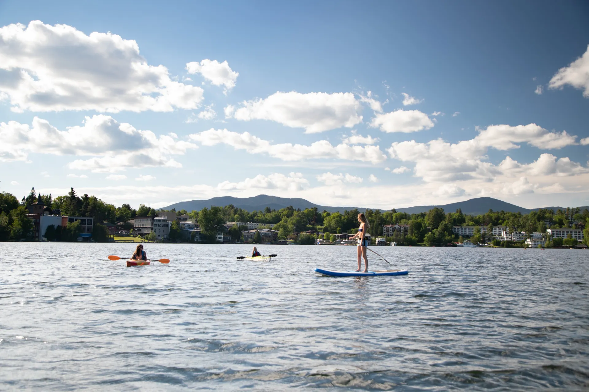 Blonde woman stands on stand up paddle board talking to two people in kayaks in middle of a lake surrounded by green mountains and homes